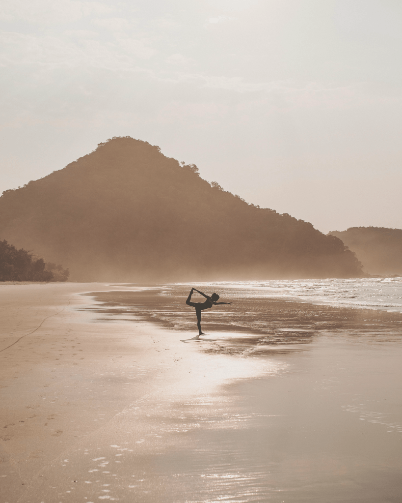 A person practising a yoga pose on a peaceful beach at sunrise, symbolising harmony between body, mind, and nature in holistic stress management.