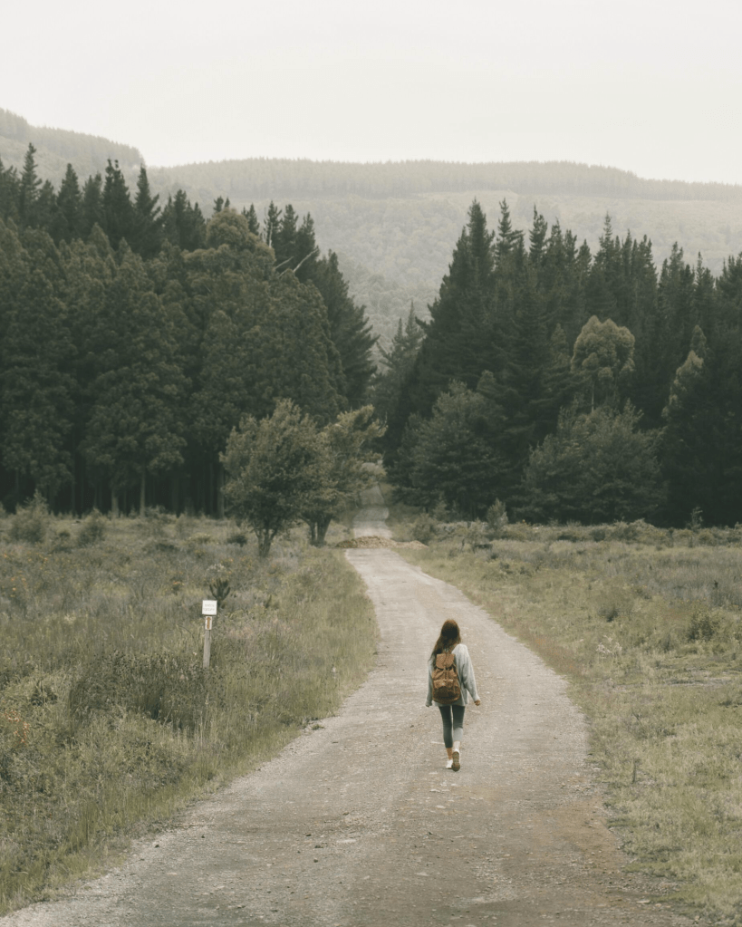A woman walking alone along a quiet forest path surrounded by trees, symbolising the personal journey of self-discovery and the courage to walk one’s own path.