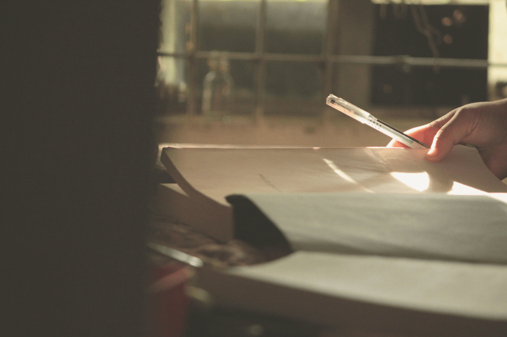 A hand holding a pen above an open notebook in soft natural light, symbolising reflection, integration, and the quiet process of self-discovery.