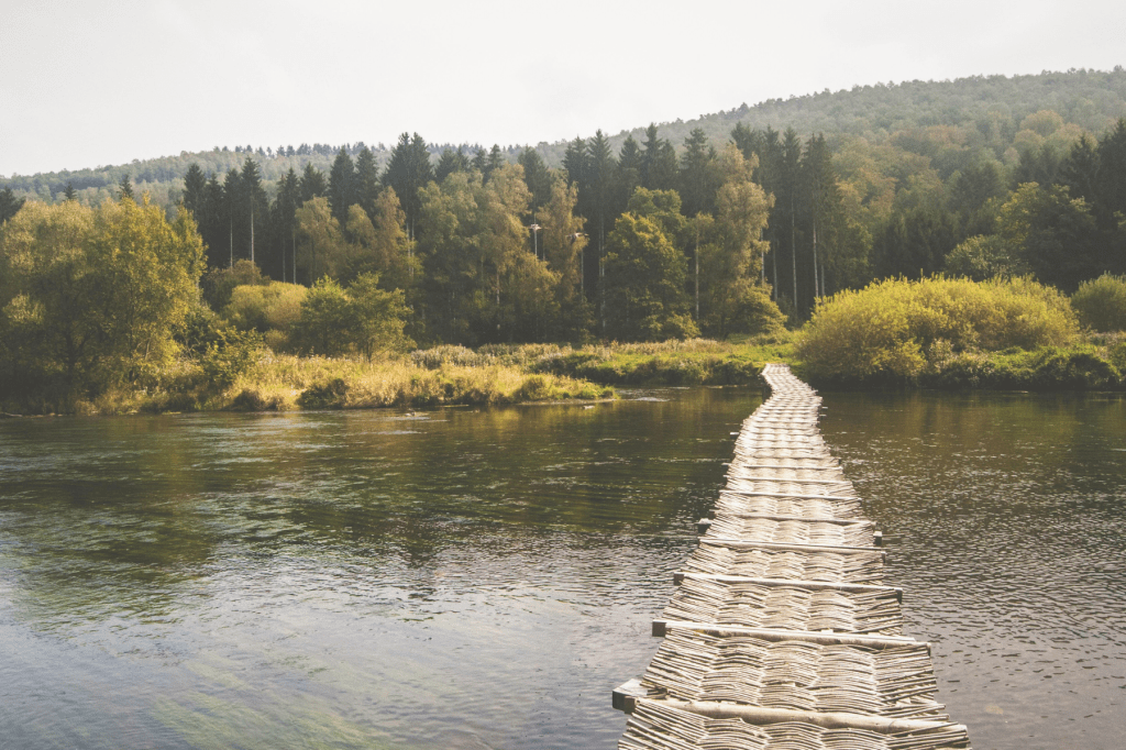 A wooden footbridge stretching across a calm river towards a forest, symbolising the ongoing journey of personal growth and self-discovery.