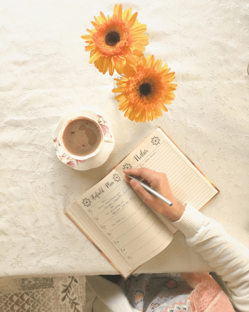 A softly lit image showing a person’s hand writing in an open notebook on a light tablecloth beside a cup of tea and yellow sunflowers. The warm, peaceful composition represents safety, reflection, and empowerment, aligning with the theme of creating a safety plan in the journey to healing from domestic abuse.