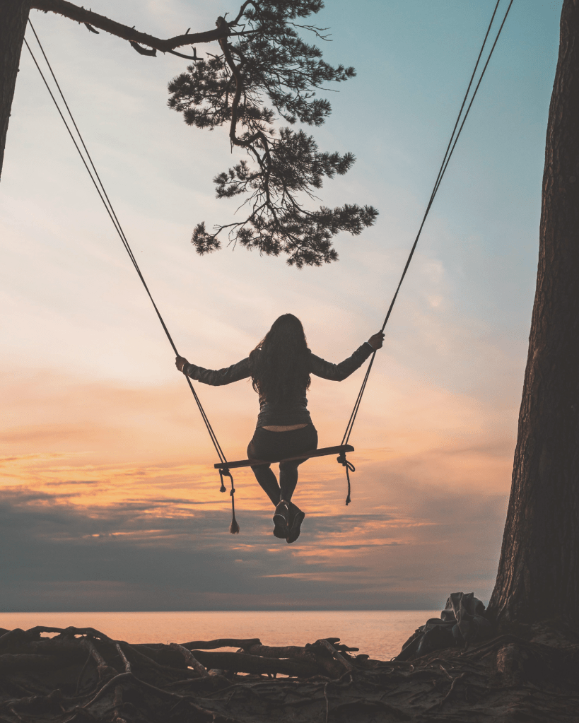 Woman on a swing overlooking a calm sunset, symbolising freedom, nervous system regulation, and the gentle rebuilding of inner safety.