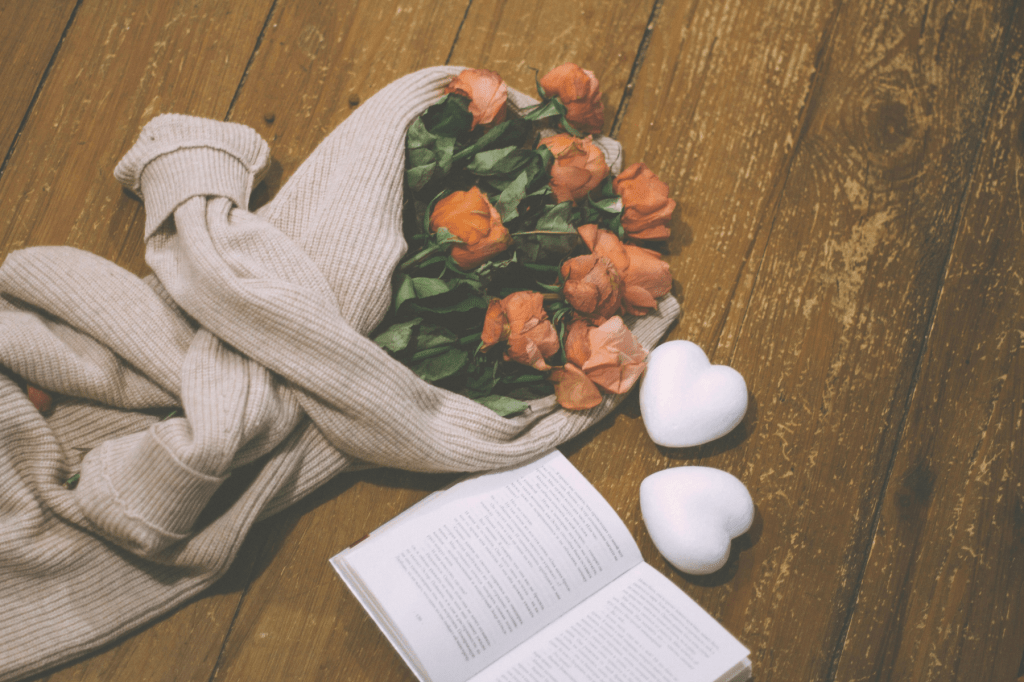 A soft folded sweater holding wilted roses beside an open book and two heart shapes on a rustic wooden surface, symbolising self compassion, healing wisdom and emotional tending.