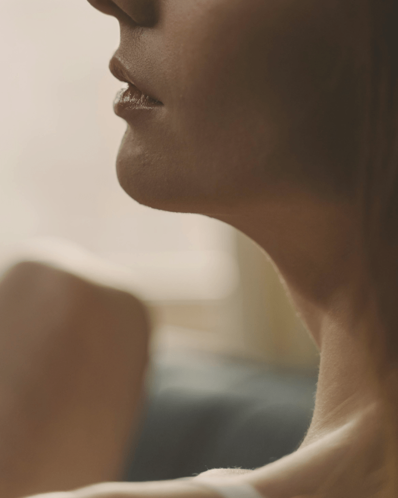 Side profile of a woman’s neck and lower face in soft natural light, with a neutral background.