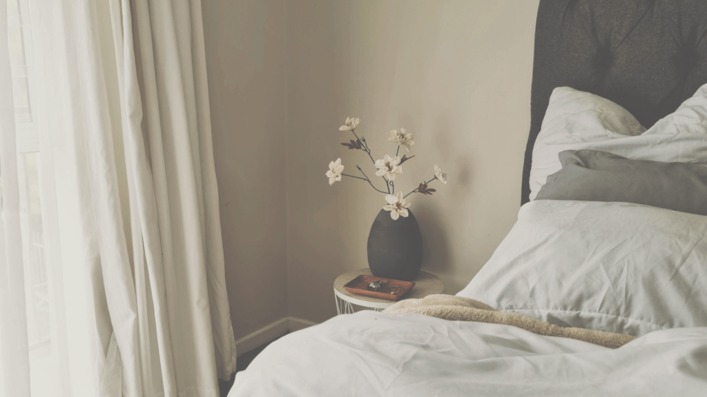 Soft natural light in a calm bedroom with linen curtains, neutral bedding and a simple vase, creating a quiet, stable interior space.