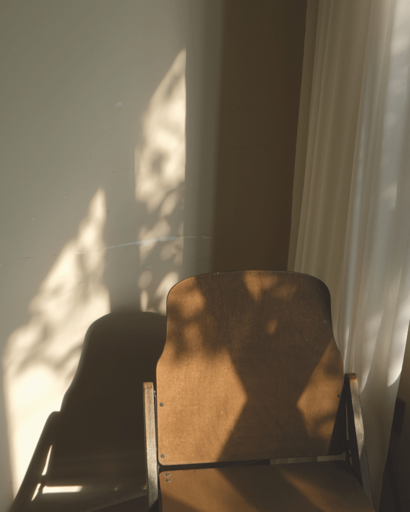 Empty chair beside soft window light and shadows, symbolising emotional space, reflection, nervous system healing and boundaries after trauma.