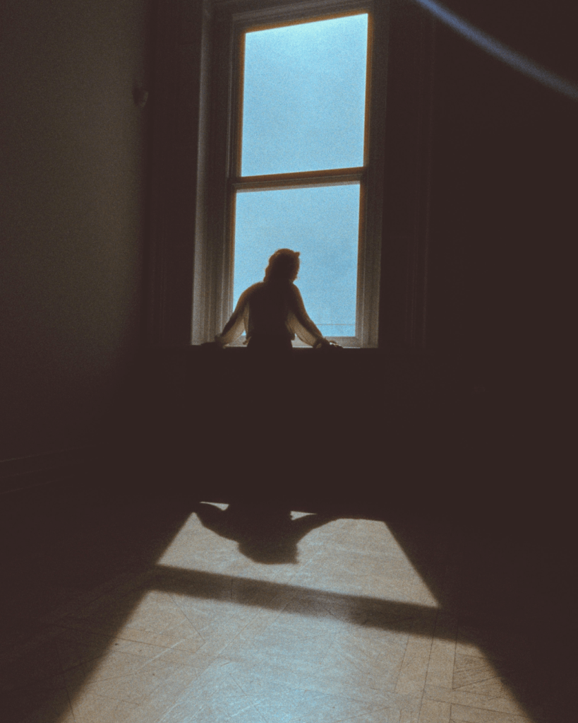 Silhouette sitting by a window in a dark room representing isolation, emotional withdrawal and identity confusion after coercive control and psychological trauma.