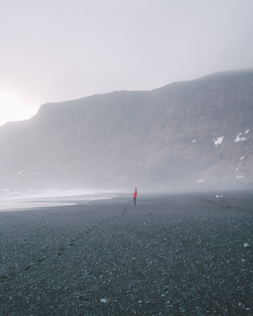Person standing alone on a misty black sand beach beneath towering cliffs, symbolising the gradual journey of trauma recovery and nervous system integration beyond intellectual insight.
